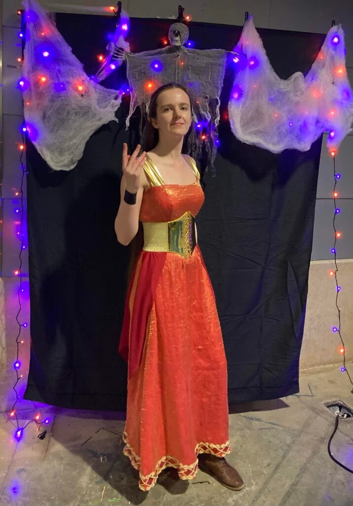 Photo of Riverly Twardy wearing a very early, incomplete version her Kyndara cosplay at a Halloween event, posing in front of a Halloween themed backdrop.