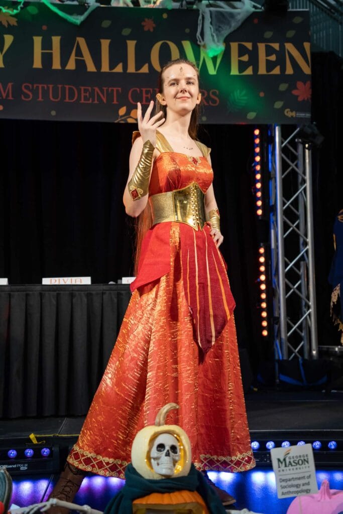 Photo of Riverly Twardy wearing a mostly complete version her Kyndara cosplay in the 2025 Halloween costume contest at George Mason University. A banner in the background has text reading "Halloween" and "Student Centers", though part of the latter is covered up.